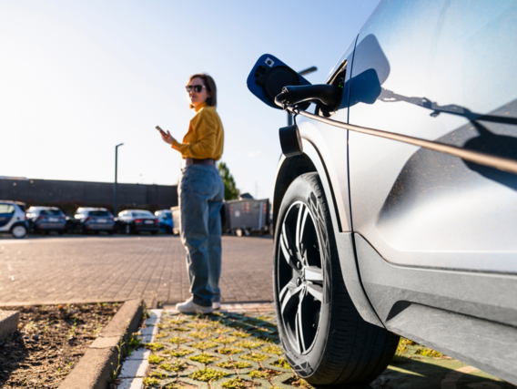 Elektroauto wird an einer Ladestation geladen, im Hintergrund steht eine Frau mit Smartphone.
