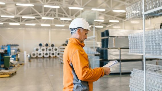 Mitarbeiter mit Schutzhelm kontrolliert Notizen in einer Produktions- oder Lagerhalle.