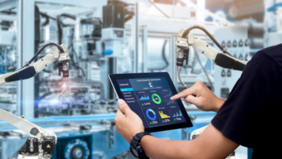 Person steuert Produktionsprozess über Tablet in automatisierter Fabrikumgebung mit Robotikarmen.
